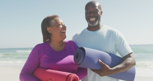 Senior couple laughing and holding yoga mats on the beach, promoting active and healthy lifestyle. Suitable for use in wellness blogs, fitness advertisements, retirement brochures, and health-related articles that focus on physical activities for mature adults.