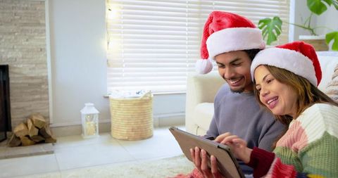 Young couple sharing tablet on living room rug wearing Santa hats and cozy sweaters