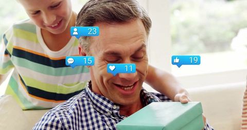 Father opening turquoise gift with child in cozy living room, social media likes overlay
