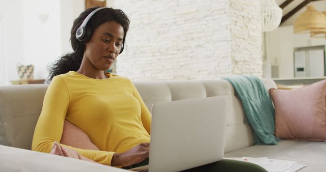 Image of happy african american woman on sofa with headphones using laptop. leisure and relax, spending free time with technology at home.