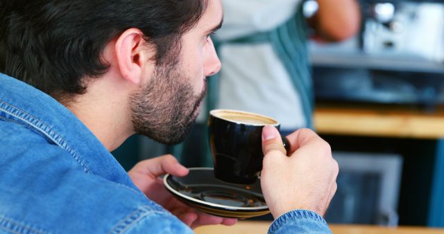 Man is drinking a cup of coffee in a cozy café while wearing a jean jacket. This scene evokes a sense of relaxation and casual lifestyle, perfect for advertising coffee products, promoting cafés, or illustrating articles about coffee culture and daily rituals.