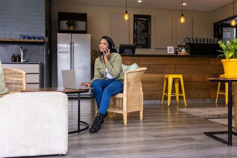 Indian woman working remotely in cozy cafe on wicker chair with laptop and phone