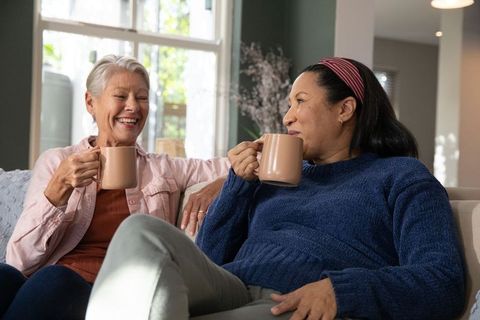 Diverse women sharing coffee and conversation on cozy sunlit sofa, laughing and relaxing