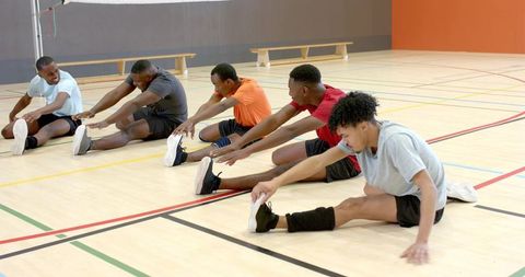 Diverse male athletes stretching and reaching toward feet on indoor gym court