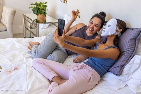 Diverse friends enjoying pamper night with led facial mask, gua sha and smartphone selfie