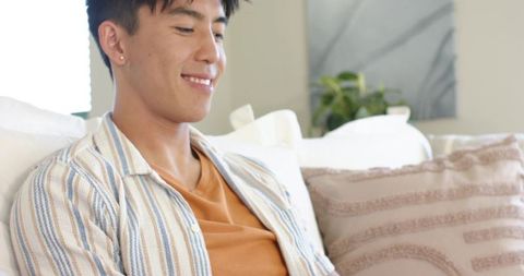Smiling asian man relaxing on sofa in sunlit modern living room with striped overshirt