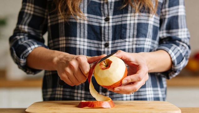 Hands peeling red apple with paring knife on wooden board in sunlit home kitchen, plaid shirt