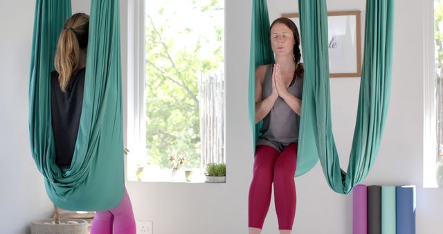 Women engaged in aerial yoga practice hanging in green hammocks in bright yoga studio. They are focused on mindfulness and relaxation. Suitable for illustrating themes of fitness, wellness, meditation, and healthy lifestyle.