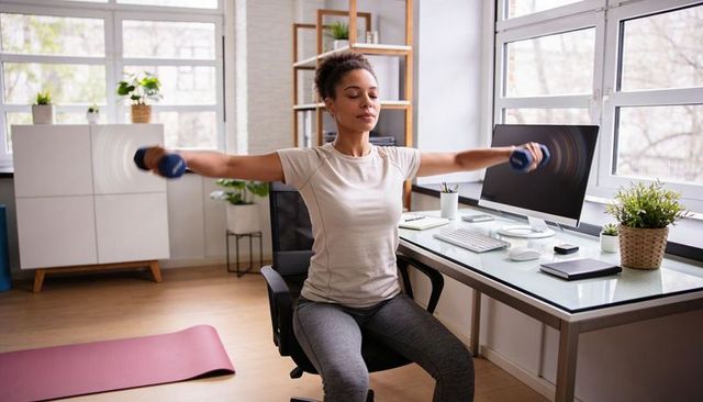 Woman exercising in home office doing seated lateral raises with dumbbells, natural light