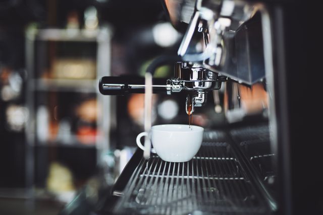 This close-up captures the moment when a professional espresso machine is pouring fresh coffee into a white cup, highlighting the detail and precision involved in making coffee at a cafe. Ideal for use in advertisements for coffee shops, promotional materials for cafes, or content related to the coffee brewing process. Great for illustrating themes of morning routines and specialty coffee preparation.