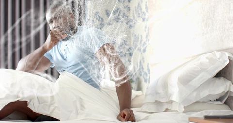 Backlit man sitting on bed touching chin holding smartphone, hazy morning contemplation
