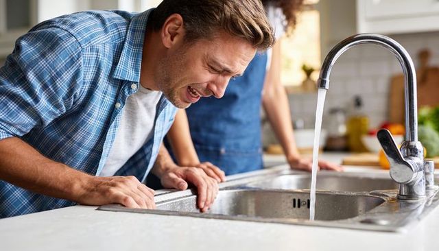 Man leaning over kitchen sink experiencing nausea with running faucet and concerned partner nearby