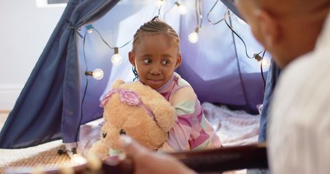African american girl hugging teddy in blue teepee with guitar and fairy lights cozy night