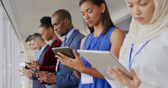 Diverse business professionals standing in line using digital tablets and smartphones. Business people in corporate attire are focusing on their devices, indicating multitasking and digitized office work. Perfect for use in articles about business technology, digital working environments, teamwork, and corporate networking events.