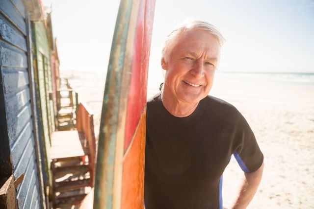 Senior man standing by surfboard at beach, smiling and enjoying a sunny day. Ideal for promoting active lifestyles, retirement activities, beach vacations, and healthy living for older adults.