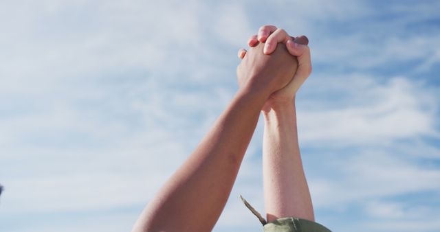 Two individuals show unity by joining hands against a clear, bright sky. Perfect for concepts of teamwork, trust, collaboration, and solidarity. Ideal for promotional materials, motivational posters, or diversity and inclusion campaigns.