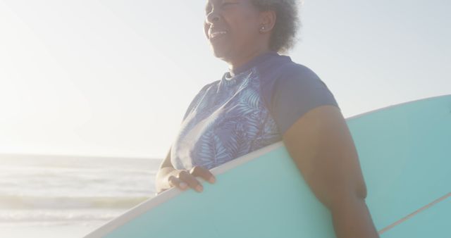 Senior woman smiling while holding surfboard on sunny beach. Great for content about active seniors, adventure, ocean sports, and enjoying life at any age.