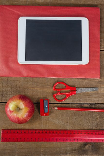 This image shows an overhead view of various school supplies including a digital tablet, an apple, red scissors, a red pencil, a red ruler, and a red sharpener on a wooden table. Ideal for use in educational content, back-to-school promotions, classroom materials, and technology in education themes.