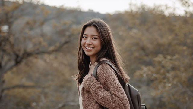Smiling student turning toward camera on autumn hillside wearing brown sweater and backpack