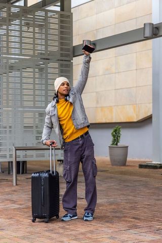 Urban traveler raising smartphone while waiting with carry-on luggage and headphones