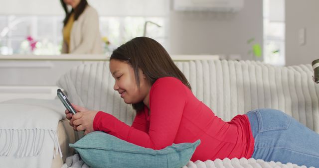 Girl is enjoying her time using a smartphone lying on a comfortable couch in a modern living room with her parent in the background. Ideal for illustrating concepts of technology use among children, relaxation, home life, or online activities.