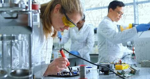 Woman soldering circuit board and testing electronics in modern lab with collaborative team