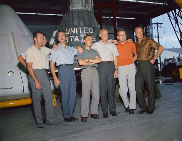 A 1963 image of the original Mercury astronauts taken at the Manned Spacecraft Center in Houston, Texas. They are standing in front of a United States spacecraft. Six men in casual attire are posing for the photo, with the spacecraft as the backdrop. Vintage images like this are useful for projects related to space history, documentaries, educational materials, and vintage photo collections.