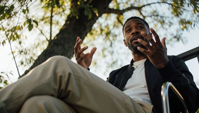 Man speaking and gesturing while sitting on sunlit park bench under leafy canopy