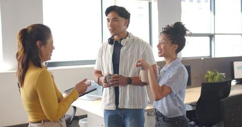 Diverse coworkers chatting and smiling in modern open office holding coffee mugs, tablet