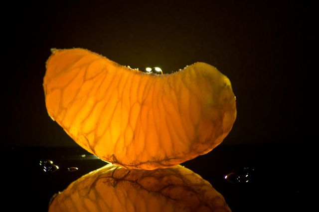 This vibrant image of a backlit orange segment captures the texture and juiciness of the fruit, making it perfect for use in food blog illustrations, health and nutrition articles, or creative advertisements for fresh produce. The dark background enhances the glowing effect of the citrus, creating a striking and eye-catching visual for various digital and print uses.