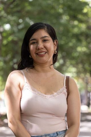 Smiling woman standing on tree-lined path wearing pink camisole, jeans and gold hoops