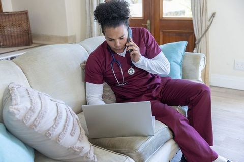 Nurse conducting telehealth call while using laptop on sofa, multitasking healthcare worker