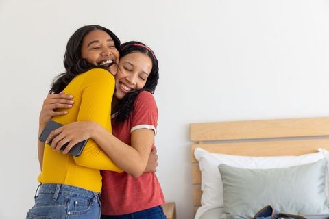 Diverse young women hugging in cozy bedroom, smiling friends embracing with smartphone
