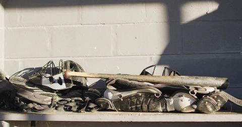 Weathered catcher's gear stacked on dugout bench with bat, masks, shin pads
