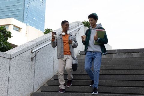 Two male students walking down urban campus stairs holding coffee, notebooks, headphones
