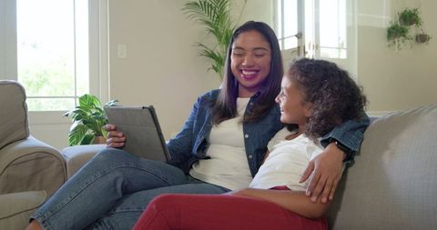 Mother and daughter sharing tablet story on sofa, smiling and bonding in bright home with plants