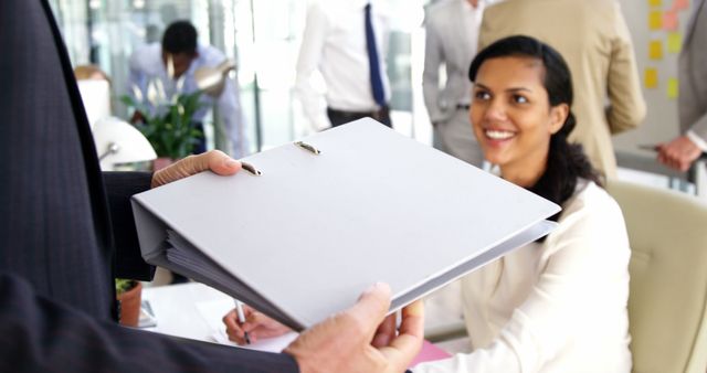 Businesswoman is sitting at desk receiving a presentation folder from colleague. Others are conversing in the background, suggesting an active, collaborative office environment. Perfect for business, teamwork, or corporate office themes.