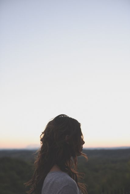 Woman with curly hair standing outdoors, facing sunrise or sunset. Silhouette set against a peaceful pastel sky, perfect for depicting contemplation, serenity, and connection with nature. Ideal for use in wellness, mindfulness, mental health awareness, solitary reflection, and inspirational projects.