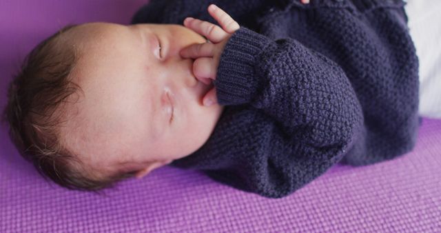 This image captures a sleeping newborn baby lying on a purple blanket with a hand resting gently on their face. The child is dressed in a cozy dark sweater, conveying a peaceful and serene atmosphere. This can be used in advertisements and articles relating to early childhood, parenting tips, nursery products, and sleep tips for newborns.