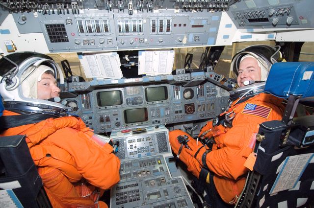 Image depicting two astronauts in orange spacesuits engaged in training session within the cockpit of a space shuttle mockup at Johnson Space Center. Can be used for content related to space exploration, NASA missions, astronaut training, and spaceship technology.