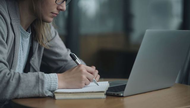 Focused woman writing notes on laptop at cafe table, remote study and productivity