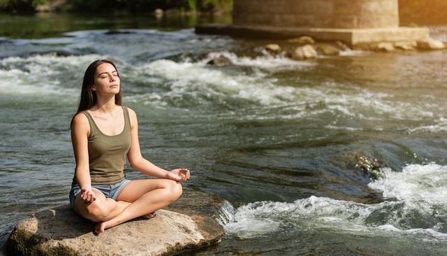 Young woman meditating on riverside rock beside flowing rapids with bridge pier and sun flare