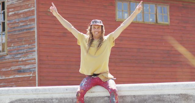 A man with long hair sitting outside against an aged red wooden building, smiling and raising both arms in a victory pose. This image can be used for concepts like joy, outside relaxation, carefree lifestyle, and celebrating successful moments.