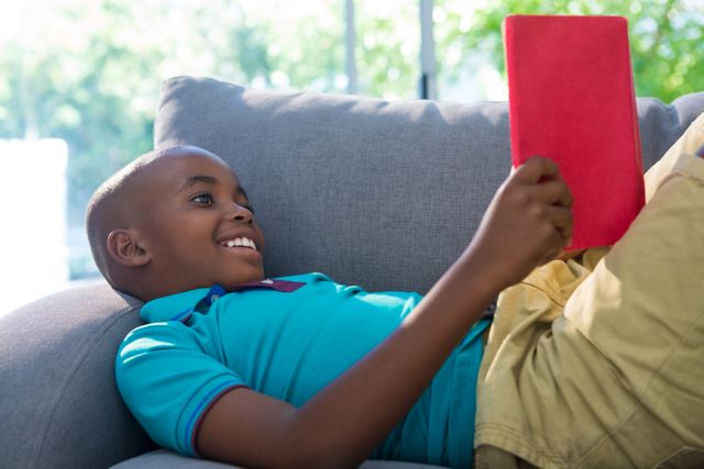 Side view of smiling boy lying while reading novel on sofa in living room at home