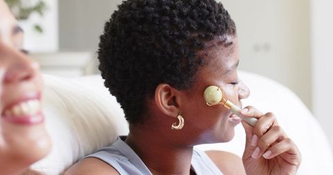 African american women doing skincare with jade roller on couch sharing relaxing self-care