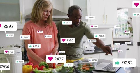 Mature couple preparing fresh salad in modern kitchen with laptop and social media likes