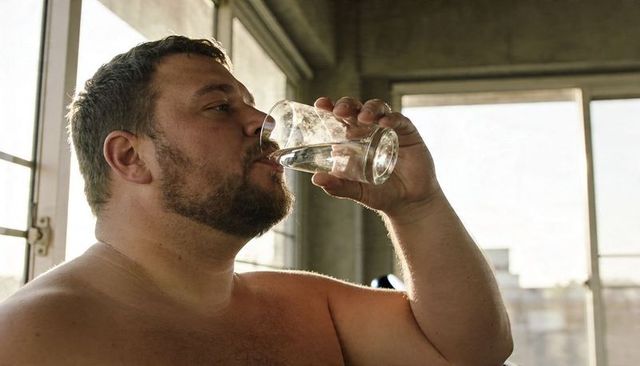 Shirtless man drinking water from clear glass tumbler in sunlit industrial loft interior