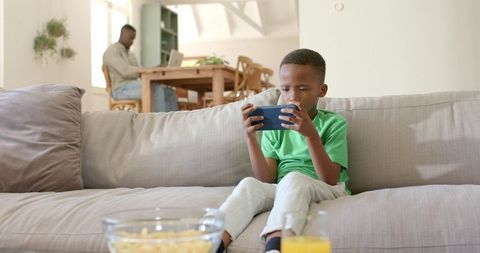 Young boy playing smartphone on sofa while father working on laptop in open living room