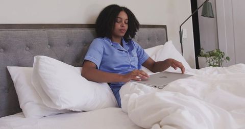 African american woman sitting in bed using laptop wearing blue pajamas minimalist bedroom
