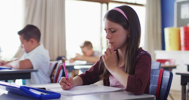 Young students are sitting at their desks in a classroom, focusing on their tasks. One girl is seen concentrating intently on her worksheet while holding a pencil. The classroom environment is active with other children also engaged in their work. This image is ideal for educational content, school advertisements, and articles related to child development and learning.
